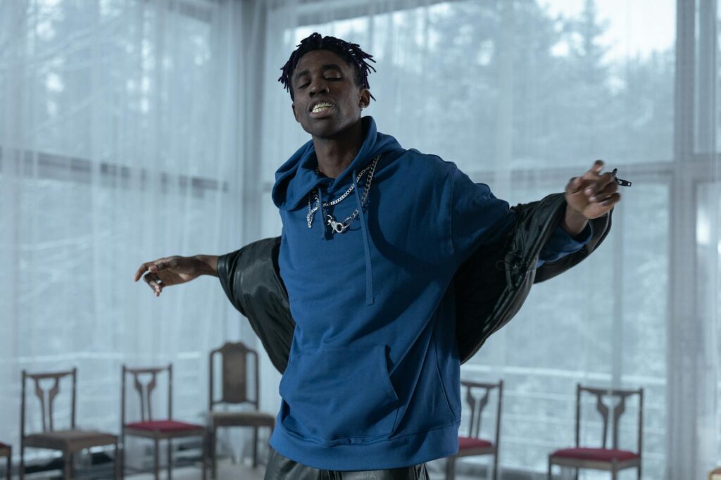 Black rapper energetically performing indoors in a blue hoodie and silver chains, conveying passion and rhythm.